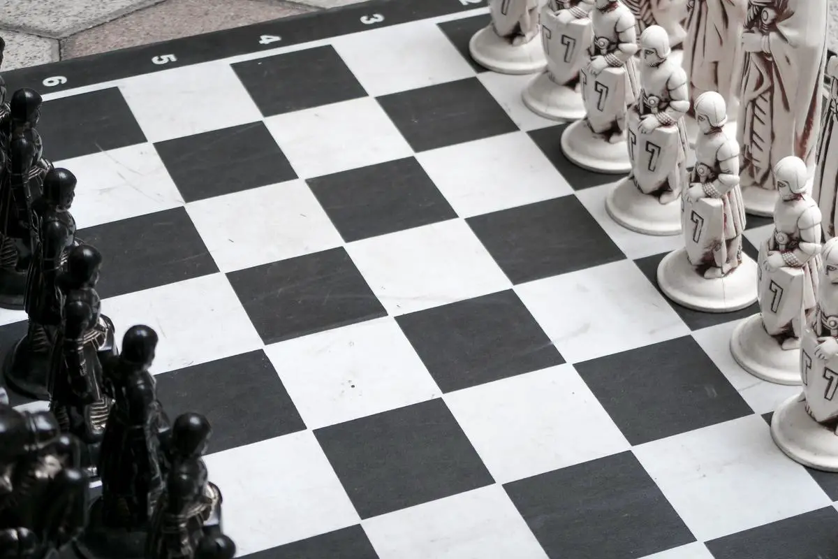A chessboard with chess pieces set up on it with two people playing and people watching behind them