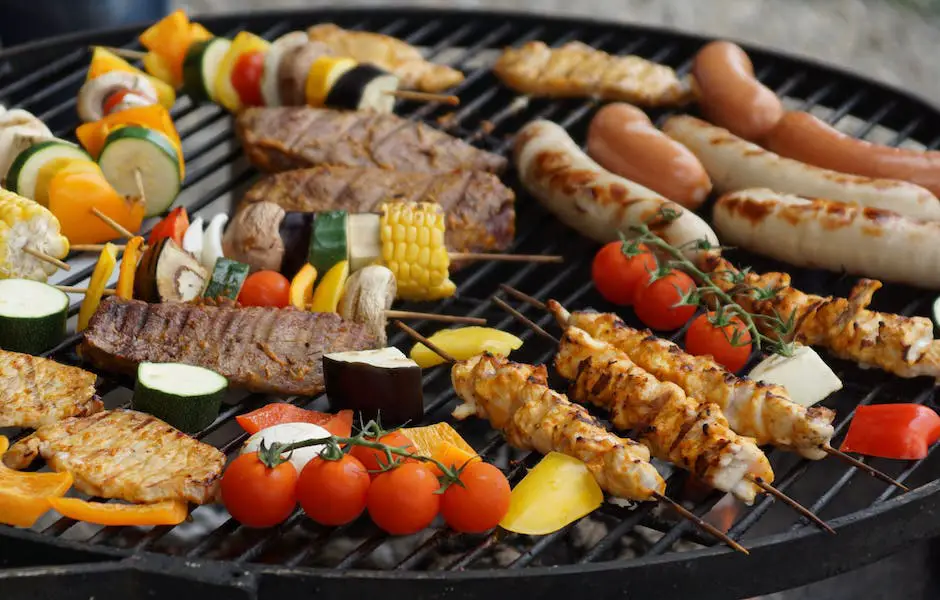 Image of a grill with burgers and vegetables cooking on it and a group of people tailgating in the background